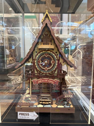 The image shows a detailed, intricate wooden model of a clock tower or a whimsical house, possibly from a storybook theme. The model is displayed behind glass, suggesting it is in a shop or museum. The house-like structure has a steep, gabled roof with ornate trim, a prominent circular clock face with Roman numerals and moving hands, and various decorative elements like hearts, leaves, and a banner that reads "Fell down the rabbit hole." At the base of the model, there's a small fence, a rabbit character holding a trumpet, and a sign that says "ROKR." A prominent sticker on the glass in the foreground reads "PRESS First step of the stairs" with an arrow pointing to the right. The background is blurred, showing shelves with books or other items, and reflections on the glass. The lighting is somewhat diffuse, highlighting the craftsmanship of the wooden model. The scene appears to be inside a retail establishment, possibly a gift shop or a specialty toy store, in Mission Viejo, United States of America.