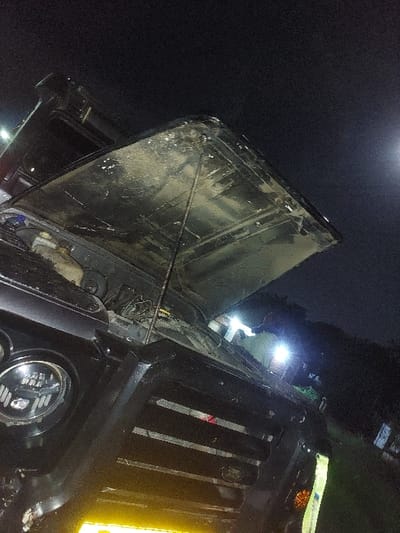 The scene depicts a dark-colored, likely black, Land Rover Defender vehicle at night in Msata, Tanzania. The vehicle's hood is open and propped upwards, exposing the engine compartment. The underside of the hood and portions of the vehicle's exterior are visibly covered in dirt and mud, indicating recent off-road travel or exposure to muddy conditions. Circular LED headlights are present on the front, along with a prominent grille featuring "Land Rover" badging. A yellow reflective strip or license plate area is visible at the bottom front, and a vertical reflective strip is affixed to the right side of the front bumper.

In the background, at least one individual is partially visible standing near the vehicle, illuminated by multiple intense white artificial light sources. These lights cast significant illumination on the person and the area behind the vehicle. The overall environment is dark, suggesting a rural or unlit area, with faint outlines of foliage or trees visible in the distance. A diffuse light source, possibly the moon or a distant street light, is discernible in the upper right portion of the dark sky. The open hood implies ongoing or impending mechanical inspection, repair, or maintenance.