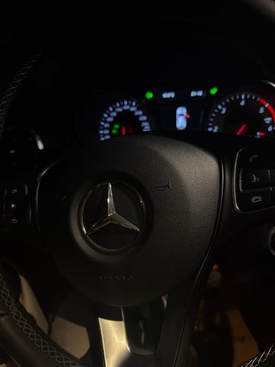 The image displays the interior of a car, specifically the steering wheel and a portion of the illuminated instrument cluster, viewed from the driver's perspective in a dark setting, suggesting night.
The central subject is a black, textured steering wheel, featuring a prominent silver Mercedes-Benz three-pointed star logo in its center. Faintly visible above the logo is the word "AIRBAG," and a small horn symbol is embossed below it. The steering wheel includes various controls: on the upper left spoke, buttons with illuminated orange symbols for picking up calls and voice control are present. On the upper right, a similar button for ending calls is visible. On the lower left spoke, there's a button marked "OK" along with navigation arrows and a menu icon. The rim of the steering wheel shows white contrast stitching.
To the left of the steering wheel, a segment of the digital instrument cluster is brightly lit. It displays white numerical readouts, red gauge needles, and green indicator lights. Blurry text on the cluster appears to show "21°C" (likely temperature), "AUTO," and a graphic depicting a car with an open door or trunk. The overall scene is dark, highlighting the illuminated car controls and displays. The car is situated in Voiniagovo, Bulgaria.
