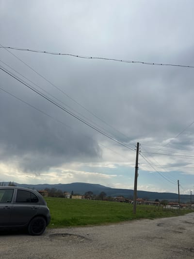 The image, taken in Padarevo, Bulgaria, captures a rural landscape under an overcast sky. The scene is viewed from a low angle, looking across a dirt road towards mountains in the distance.
In the foreground on the right, a dark gray hatchback car is parked on the unpaved road. The car has black wheels and what appear to be tinted windows. The road itself is rough, with visible stones and patches of bare earth.
A wide expanse of vibrant green grass or a field occupies the middle ground, stretching from the road towards a distant mountain range. Several wooden utility poles with numerous power and communication lines traverse the scene. One prominent pole stands on the right side of the field, extending upwards into the sky. Other power lines run diagonally across the frame, particularly from the upper left.
In the background, a range of dark, undulating mountains rises, partially shrouded in haze. Below the mountains, scattered houses with red-tiled roofs and other structures are visible, indicating a village or settlement nestled in the valley. The sky is predominantly covered in a thick layer of gray clouds, with lighter, whiter clouds appearing towards the right, suggesting dynamic weather patterns or a break in the overcast conditions. The scene appears to be daytime, likely an overcast day. No specific activity or visible text is present.