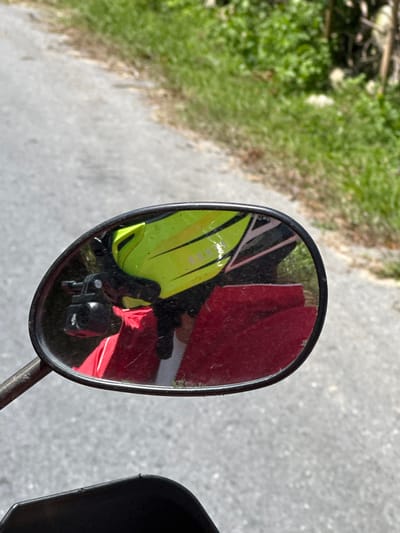 A close-up shot depicts a motorcycle mirror reflecting a scene. A person wearing a yellow and black high-visibility vest is seen. Also reflected is a red object. The surrounding environment appears to be a paved area next to a grassy verge. This image may have been taken in the vicinity of Bản Hồ, Vietnam.