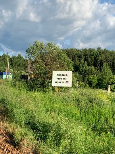 A white sign with black text is situated in a field of tall green grass and wildflowers. The sign reads "Хорошо, что ты приехал!!!" which translates to "It's good that you've arrived!!!". In the background, a dense forest of pine and deciduous trees forms the horizon. To the left of the sign, a small, weathered wooden structure with a blue roof is partially visible, along with a utility pole. The sky above is dramatic, with a mix of dark, heavy clouds and patches of bright blue. The overall scene suggests a rural or semi-rural setting, possibly an entrance to a village or a remote property, given the presence of the sign and the rustic buildings. The location is identified as Svetitsa, Russia.