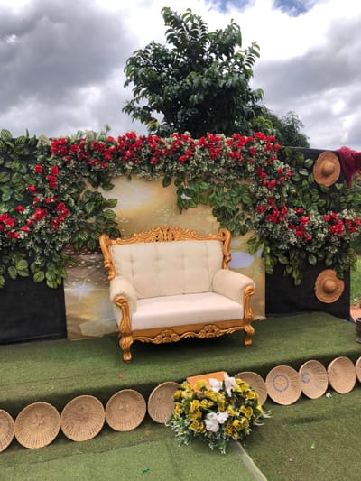 The image depicts an outdoor event setup in Amanuke, Nigeria, under an overcast sky. A cream-colored two-seater sofa, featuring an ornate gold-painted wooden frame and button-tufted upholstery, is centered on a raised platform covered in artificial green turf. Behind the sofa, a backdrop is arranged, consisting of a central light-colored panel with a blurred design, flanked by two black panels. Artificial green foliage and numerous red roses are draped across the top and down the sides of the backdrop, forming a floral arch. Two circular woven straw hats are affixed to the right black panel, with a piece of dark red fabric draped nearby. In the foreground, on the green turf, a row of approximately ten circular, light-brown woven mats or shallow baskets is present. A bouquet of artificial yellow and white flowers with green foliage sits on the turf to the right of these mats, partially obscuring a small, light-colored rectangular object. A large green tree is visible in the background, above the floral display. No individuals are present.