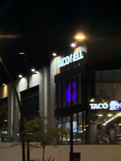 The image shows a nighttime view of a Taco Bell restaurant, likely in the city of Alcorcón, Spain, as indicated by the prompt. The building features prominent white neon signage with the "TACO BELL" logo and a glowing purple Taco Bell bell icon. Several exterior lights illuminate the facade of the building. There are large windows through which some interior lighting and reflections are visible. The scene appears to be captured from a moving vehicle, as indicated by the slight blur and motion in the image, particularly noticeable in the foreground elements like trees and streetlights. The overall environment is dark, typical of nighttime in an urban setting, with artificial light sources being the primary illumination.