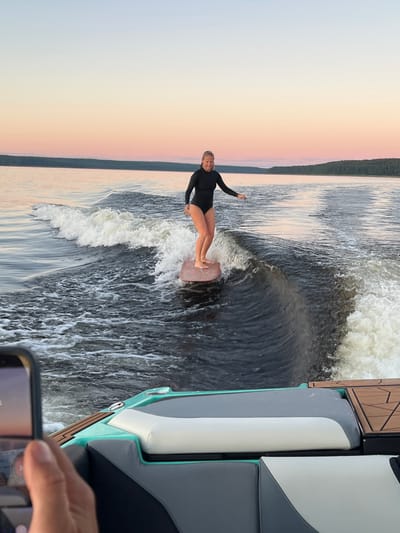A woman in a black swimsuit is wake surfing on a calm lake during sunset in Gromovo, Russia. She is standing on a pink surfboard, riding a wave generated by a boat. The sky is a gradient of soft pink, orange, and blue. In the foreground, a hand holds a smartphone, capturing the scene. The boat's seating area, with grey and white upholstery, is partially visible. The environment is serene and picturesque, with a treeline on the horizon.
