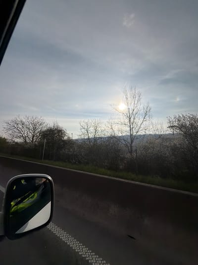 A daytime shot taken from inside a moving vehicle shows an overcast sky with the sun visible through bare tree branches. Outside the window, vegetation, likely trees, covers a slope leading to what appears to be a residential area, possibly located in the city of Mertert, Luxembourg, although the exact location cannot be confirmed. The side mirror of the vehicle reflects the driver wearing a yellow vest. A road with lane markings is visible in the lower right quadrant of the frame.