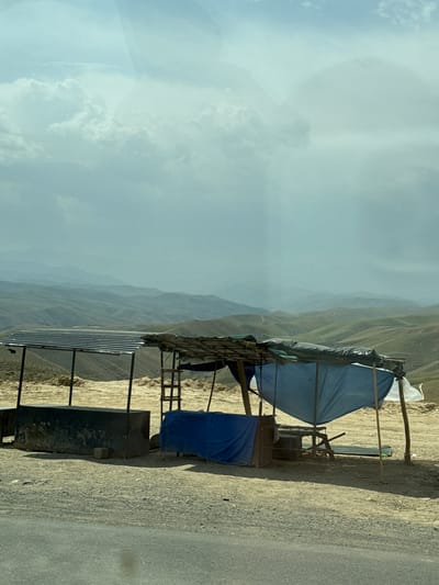 The image depicts a roadside stall in a mountainous, arid region. The stall is constructed from wooden poles and covered with tarps, one blue and one white, providing shade from the sun. Beneath the tarps, there are various objects, including what appears to be a metal container or structure and a corrugated metal sheeting structure. The ground is covered with gravel and sand, characteristic of a dry environment.

In the background, rolling hills and mountains recede into the distance, shrouded in a hazy atmosphere. The sky is partly cloudy, with large, white cumulus clouds dominating the upper portion of the image, suggesting fair weather. The overall setting appears remote and desolate, with the road running alongside the stall.

The image is oriented vertically, with the sky on the left and the ground and stall on the right. There is no visible text in the image. The time of day appears to be daytime, likely late morning or early afternoon, given the lighting conditions. The scene conveys a sense of a temporary or makeshift structure set up to serve travelers in a sparsely populated area. The location context suggests Baytepa, Uzbekistan.
