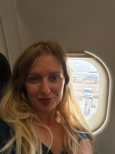 A woman with long blonde hair is seated on an airplane, looking at the camera. She is wearing a dark blue shirt. Through the airplane window, an Arkia airplane is visible on the tarmac, along with service vehicles. The scene appears to be at Ben Gurion Airport in Yehud-Monosson, Israel. The lighting suggests it is daytime.