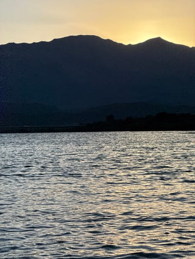 A landscape photograph, taken in Dumbéa, France, depicts a natural waterscape under low light conditions, indicative of either dawn or dusk. The lower two-thirds of the image are occupied by a body of water with a textured surface of small ripples, reflecting pale yellow and grey light. A thin, dark, indistinct landmass or distant shoreline is visible at the transition from the water. The middle ground features a prominent, dark, and irregularly shaped mountain range presented in silhouette, with its peaks and valleys defining the horizon. The upper third of the image consists of a sky with a concentrated yellow-orange glow along the mountain crests, suggesting the presence of the sun just below the visible frame, transitioning to a lighter, more muted tone overhead. No human subjects, artificial structures, or explicit actions are present within the frame.