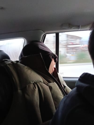 The scene is set inside a moving vehicle in Dragievtsi, Bulgaria. A person is seated in the rear passenger compartment, positioned in profile looking out the right-side window. This individual wears a dark hooded garment with the hood pulled up, partially obscuring their face, and an olive-green quilted puffer vest or jacket over it. A segment of their light-toned chin and mouth area is discernible. In the immediate foreground, to the right, the out-of-focus dark shoulder and a partial view of the head of another occupant are visible. Through the vehicle's side window, a blurred exterior environment is observable, featuring indistinct buildings and distant terrain, consistent with motion through a populated area.