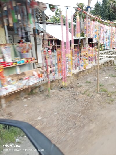 The image shows a street vendor selling fireworks in Kecamatan Gabuswetan, Indonesia. The vendor has arranged the fireworks on a wooden stand and has hung them on bamboo poles. The fireworks are colorful and diverse, with different sizes and shapes. The vendor's stand is located on the side of a dirt road. The road is covered with gravel and is dusty. There are no visible people in the image. The image is blurry, suggesting it was taken while in motion. The image is taken from a low angle, likely a moving vehicle. The date and time displayed on the image are March 6, 2025, at 17:05.
