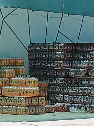 The image shows a close-up of stacks of canned beverages, likely in a market or storage area. The cans are arranged in rows and columns, with some stacks reaching higher than others. The background is blurred, but appears to be an outdoor setting with a teal-colored awning or covering supported by dark poles. The overall impression is of a commercial or retail environment. The image is too low resolution to determine specific brands of beverages or the exact location within Gulistan, Uzbekistan.
