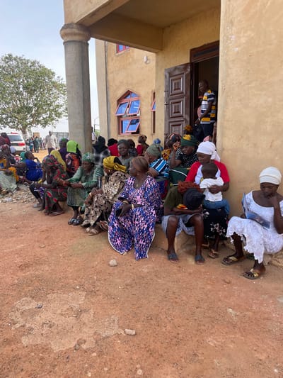 A large group of people, predominantly women and children, are gathered and seated on the reddish-brown, unpaved ground outside a light-colored building in Rahwol Chom, Nigeria. The scene appears to be a community gathering or event, taking place during the daytime under clear, sunny skies.
Many of the women are dressed in vibrant, patterned traditional attire and wear head wraps. They are seated closely together in rows, facing towards the right side of the frame, suggesting they are observing or listening to something out of view. One woman in a white dress and head wrap, located towards the upper right, is seen reading from a book or pamphlet. Another woman towards the center holds a young child on her lap. A few men are visible, including one in a striped shirt standing in the doorway of the building.
The building itself has a light tan-colored exterior with a wooden door featuring carved panels and several open, blue-tinted windows. A sturdy concrete pillar is visible on the far left, suggesting a covered porch or structural support. In the background, a large tree with green foliage stands against the bright sky, and outlines of other buildings and vehicles can be faintly seen, indicating a populated area. The ground shows patches of compacted earth and some loose stones. No legible text is visible in the image.