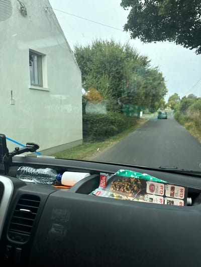 The image is a view from inside a vehicle, looking out through the windshield. In the foreground, on the dashboard, are a bottle of water, a roll of paper towels, and some folded leaflets or a flyer with pictures of food and prices. To the left, a car mount with a suction cup is visible, holding what appears to be a blue object. The vehicle is on a paved road that curves to the right. On the left side of the road is a white house with a window and some exterior wiring. On the right side of the road are dense green trees and foliage. Further down the road, another car is visible. The sky is overcast. The scene is likely set in Bamford Cross, Ireland, given the context.