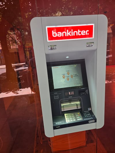 An outdoor Bankinter ATM is shown, its screen displaying a map with several location markers and the text "Y allá.", "Y allí.", "Y aquí.", and "Y aquí también.". Below the screen are a card slot, a keypad, and slots for bills and receipts. The ATM is situated behind a reflective surface, likely a window, which shows a blurred reflection of trees and buildings, including signage for "PLAZA". The overall environment suggests an urban setting, possibly a street-facing location in Collado Villalba, Spain, given the Bankinter branding. The lighting is bright, indicating daytime. There are no visible people directly interacting with the ATM, though a faint reflection suggests a person is present near the reflective surface.