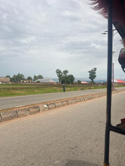 A wide shot shows a man standing on the side of a road in Kona Garu, Nigeria, with a cloudy sky overhead. The man, wearing a dark shirt and blue jeans, stands near a concrete curb. In the background, there are several buildings with red and gray roofs, surrounded by green grass and trees. A light pole with a solar panel is visible on the right side of the image. The foreground is dominated by the asphalt road and a portion of a vehicle, likely a tricycle, suggesting the photo was taken from inside it. The overall scene depicts a rural or semi-urban environment on a cloudy day.