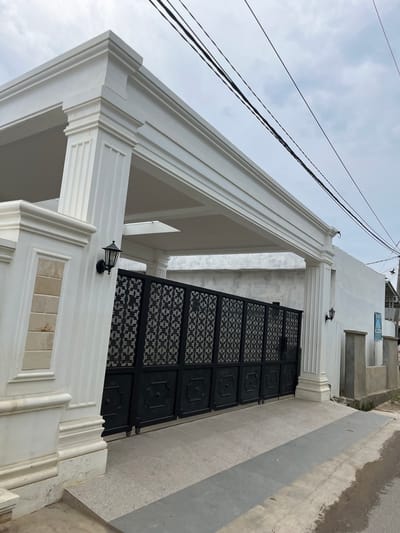 The image depicts a modern white gate and facade in Tanahterban, Indonesia. The gate is black with an intricate geometric pattern and is flanked by large white pillars. A white awning with decorative molding extends over the entrance. An outdoor lamp is mounted on the left pillar. In the background, a white wall runs alongside the property, with a street and sidewalk visible in the foreground. Overhead, black power lines are strung against a cloudy sky. The overall impression is one of a well-maintained, upscale residence.