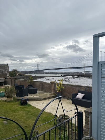 The image presents an overcast waterfront scene captured from a residential property in North Queensferry, United Kingdom. The sky is uniformly covered with dense, grey clouds, indicating diffuse light conditions.

In the background, the Firth of Forth stretches across the frame, spanned by two distinct bridges. To the left, the Forth Road Bridge, a grey suspension bridge with visible lattice work and road deck, is prominent. To its right and slightly behind, the Queensferry Crossing, a white cable-stayed bridge with slender towers, is also visible. The water surface appears calm, and a rocky, exposed foreshore, suggesting low tide, is present in the mid-distance. A traditional stone house with a dark roof is partially visible on the distant left shoreline, nestled amongst sparse vegetation.

The mid-ground features a paved patio constructed from light-colored flagstones, bordered by a low brick wall that separates it from the water's edge. On the patio, black rattan-style outdoor furniture is arranged, consisting of two armchairs, a square ottoman, and a two-seater sofa. The sofa has one decorative cushion, white with a pink geometric pattern. A small tree with variegated yellow-green leaves is contained within a dark grey pot on the patio.

The foreground consists of a neatly maintained green lawn. A decorative black wrought-iron railing with ornate scrollwork partially frames the bottom section of the image. Various potted plants, including a teal ceramic pot and other terracotta containers, are placed on the grass and near the patio's edge. On the far right, a vertical light-blue wooden structure, resembling a louvered panel or shutter, frames the scene's right boundary. No individuals are present in the depicted scene.