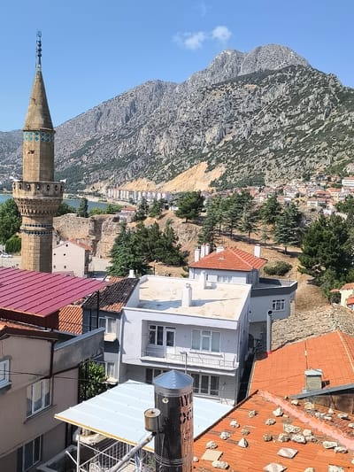 The image captures a view of Eğirdir, Turkey, dominated by a towering minaret in the foreground left. The minaret, constructed from tan-colored stone, features decorative bands of turquoise and green tiles and multiple balconies, each with what appear to be loudspeakers. Behind the minaret, a body of water, likely a lake, is visible, bordered by a town built on a hillside. This town consists of numerous buildings with red-tiled roofs, interspersed with trees and a prominent, rugged mountain range in the background. The foreground of the image is filled with the rooftops of several houses, varying in color and material from red tile to white metal. The overall scene is one of a sun-drenched day, with a clear blue sky above.