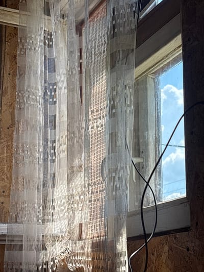 The image captures a view from under a skylight, looking upwards. The skylight frame appears to be made of wood and is set into a ceiling constructed of particle board. The glass of the skylight is somewhat dirty, with a few thin, dark wires running across its surface. Outside, a bright blue sky with fluffy white clouds is visible, suggesting it is daytime.
Dominating the foreground is a series of draped sheer curtains. These curtains have a patterned weave, creating a textured appearance with alternating bands of dense and open fabric, forming geometric shapes. The light filtering through the skylight and the curtains casts shadows and highlights, adding depth to the scene.
The overall impression is one of an unfinished or under-construction interior space. The exposed particle board ceiling and the utilitarian nature of the wiring and skylight frame suggest a functional rather than decorative setting. The presence of the curtains implies an attempt to soften the harshness of the interior or to control the light. There is no visible text in the image. The location context of Yanevo, Russia, does not provide specific visual cues within the image itself.