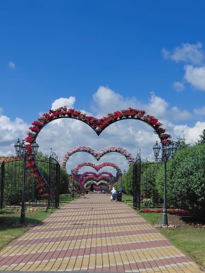 The image captures a path in a park in Kovalivka, Ukraine, adorned with a series of heart-shaped floral arches. The path is paved with alternating red and yellow bricks, creating a visually appealing pattern. On either side of the path, there are green hedges and black metal fences. Ornate black lampposts are strategically placed along the path.

The arches are densely decorated with colorful flowers, primarily red and pink, arranged in large heart shapes. The largest arch is in the foreground, framing the view of subsequent arches that recede into the distance. The sky above is a vibrant blue with scattered white clouds.

In the background, a few individuals are visible. One person is seated on a bench near a lamppost, and another is riding a blue and white bicycle. The overall scene suggests a well-maintained and aesthetically pleasing park environment, likely a popular tourist destination.