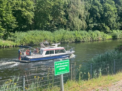An overhead view shows a body of water in Teltow, Germany, with a boat navigating the river. The boat is blue and white and has "SCHIFFAHRTSPOLIZEI" and "CHARLOTTENBURG" written on its side. A German flag is displayed on the back of the boat. On the shore, a green sign reads "Bitte die Wege nicht verlassen und die Böschungsanlagen nicht betreten!" and "REHA-ZENTRUM TELTOW". The riverbanks are lined with lush green trees and foliage, with a metal fence separating the water from a grassy area. The sky is clear and blue.