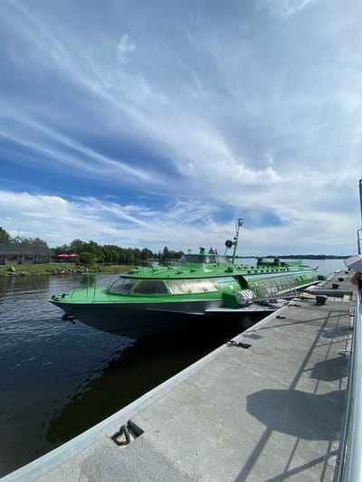 A bright green hydrofoil boat is docked at a concrete pier on a sunny day. The boat, which appears to be a passenger vessel, is positioned alongside the pier, with its bow facing left and its stern extending to the right. The water is calm and dark, reflecting the blue sky and clouds. In the background, across the water, is a treeline and a building with a red roof, suggesting a waterfront area. The pier itself is made of concrete and has railings. The sky is a vibrant blue with wispy white clouds, indicating fair weather. The scene is static, with no visible activity. This image appears to be taken in Yamka, Russia, a location known for its waterways and recreational activities.