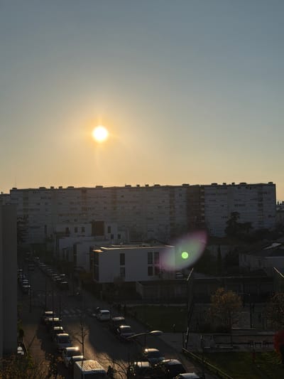 An elevated long shot captures a residential urban landscape in Bron, France, under a low-angle sun. The sun, a brilliant white orb with radiating light, is positioned in the upper left quadrant of a pale blue sky, casting a strong lens flare, visible as a green and magenta oval, in the lower right of the frame. The horizon is dominated by a long, multi-story, grey residential building featuring numerous windows, extending across the mid-ground. Below this structure, several smaller, modern white buildings with flat roofs are visible.
A street runs diagonally from the lower left towards the lower right, lined on both sides with parked vehicles, including sedans, hatchbacks, and a white van in the foreground. Sunlight reflects off the roofs and windshields of some vehicles, particularly on the left side of the street, creating bright glints. Long shadows are cast across the pavement. Leafless trees are interspersed along the street and in surrounding green spaces, with some showing sparse foliage. One individual is discernible walking on the left sidewalk, casting a long shadow, and another figure is possibly visible near the white van. Streetlights are present along the road.