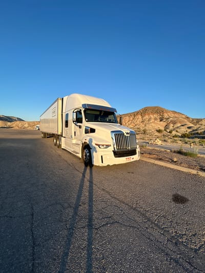 A white semi-trailer truck is parked on a cracked asphalt lot in a desert landscape. The truck has a long white trailer with "Premier" printed on the side in black lettering. The truck cab is white with a chrome grille and the Western Star logo. The sun is low in the sky, casting long shadows from the truck across the pavement. In the background, there are rolling hills with sparse vegetation and a clear blue sky. A second, smaller white truck is visible in the distance. The scene appears to be a rest stop or parking area in a rural location. There are no people visible in the image. The weather appears to be clear and sunny, likely in the late afternoon or early morning due to the long shadows and warm light.