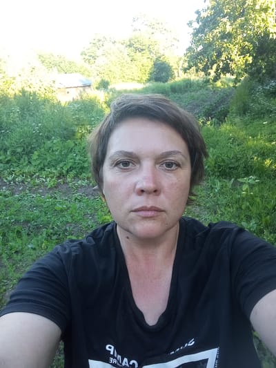 A selfie taken outdoors shows a woman in her late 30s or early 40s with short brown hair, looking directly at the camera. She is wearing a black t-shirt. Her face is centered in the frame, with the background showing lush green grass, foliage, and trees. A small wooden structure with a metal roof is visible in the distance, to the left of the woman. The overall setting appears to be a rural or semi-rural area, possibly a backyard or a countryside location. The lighting suggests it is daytime, likely late afternoon due to the warm tones and shadows. The woman has a neutral expression. The location is identified as Shil'tsevo, Russia.
