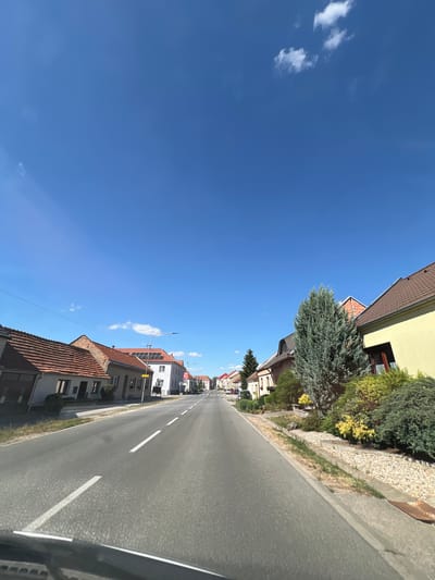 The image depicts a street scene in Kostice, Czechia, on a bright, sunny day. The perspective is from a vehicle moving along an asphalt road, which occupies the right side of the frame. White dashed lines mark the lanes of the road.
On the left side of the road, a row of houses with tiled roofs lines the street. Some houses are single-story, while others appear to be two-story buildings. Various trees and bushes, including evergreen varieties, are planted along the property lines, adding greenery to the scene. One house prominently features solar panels on its roof, suggesting a modern touch amidst the traditional architecture.
The sky is a vibrant, clear blue, with a few wispy white clouds scattered across it. The bright sunlight indicates it is likely midday. The overall atmosphere is peaceful and serene, typical of a small town or village. No people are visible in the image. The image is shot vertically, giving a dynamic perspective of the street receding into the distance.