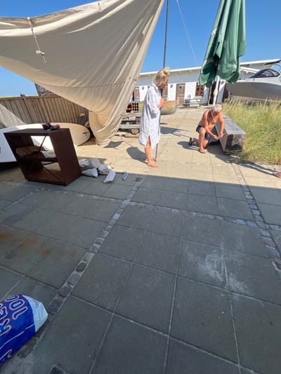 The scene depicts an outdoor paved area, likely a patio or dockside, in Skagen, Denmark, under clear sunny skies. Two individuals are present: a blonde female in a light-colored long shirt and sandals stands upright, looking towards a male. The male, with grey hair, is kneeling on the paving, attending to a large, rectangular wooden beam or bench positioned next to tall green grass. He appears to be manipulating a small, light-colored object with both hands near the end of the beam.

A large, light beige canvas tarp, supported by a pole, extends from the left foreground towards the center, casting a shadow across the lower portion of the image. To the left, a dark brown, multi-tiered shelf unit lies horizontally on the paved surface, with a red power drill resting on its top surface and several white sacks, possibly containing sand or cement, piled nearby. Two small silver cylindrical objects are also visible on the pavement near the sacks. Further left, partially visible white boards, possibly surfboards or paddleboards, lean against a wooden fence.

In the background, a white building with multiple dark wooden doors is visible, with a small outdoor table and chairs in front of it. A folded green patio umbrella stands upright to the right of the canvas tarp. Beyond the building and grass, a large white boat, resembling a yacht or cruiser, is partially visible, indicating a waterfront or harbor location. The paved ground consists of large grey square tiles bordered by smaller cobblestones, with a blue and white bag resting in the lower left foreground.