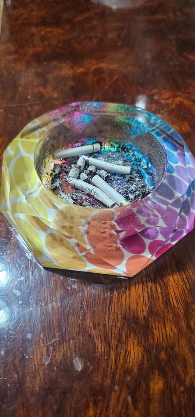 The image shows an ashtray filled with cigarette butts and ash. The ashtray is octagonal and made of clear resin with a colorful mosaic pattern of circles in shades of yellow, orange, pink, and purple. The ashtray is resting on a polished wooden surface with visible grain and some smudges or water spots. Several cigarette butts are in the ashtray, some with filters, and ash is accumulated within the bowl. One cigarette butt has visible text that reads "SOBRANIE". The lighting suggests it is an indoor setting, likely a table. The location of Novxanı, Azerbaijan, is not depicted or suggested by any visual elements in the image.
