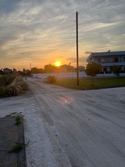 The image depicts a street scene at sunset in Chaidefto, Greece. The sky is filled with wispy clouds, tinged with the warm hues of the setting sun. A dirt road, marked with tire tracks, runs through the foreground. On the left side of the road, overgrown vegetation and some structures are visible, including a person walking away from the camera. To the right, a modern, multi-story white house with dark windows stands on a neatly mowed lawn. A single wooden utility pole with electrical wires stretches across the frame. A trash bin is positioned on the side of the road near the house. The overall impression is one of a residential area experiencing the end of the day.
