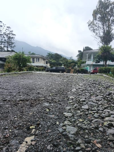 The image captures an outdoor scene in Tapaiteng Uesa, Bhutan, under an overcast sky. The foreground is dominated by a wide, unpaved surface comprised of dark gray and black gravel and larger stones, exhibiting damp patches and scattered organic debris. In the midground, a two-story building with a light-colored facade and white-framed windows is visible on the left, partially surrounded by vegetation. A dark-colored pickup truck with a bed cap is parked in front of this structure, oriented towards the right. To the right of the truck, a set of outdoor stairs ascends, flanked by low walls and decorative white, dome-like structures on pedestals. Further right, a second two-story building with a white and light blue/green exterior features a balcony supported by white columns. A red minivan is parked in front of this second building, facing left. The background consists of mountainous terrain obscured by heavy cloud cover, with dense tree lines visible on the accessible slopes. No people are present.