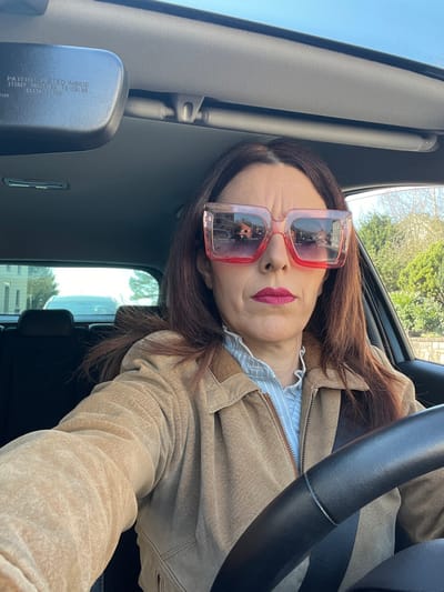 The image depicts a female individual occupying the driver's seat of a vehicle, positioned on a paved road within Roodt-sur-Syre, Luxembourg, on a clear, sunny day.
The subject is a mature female with shoulder-length, dark brown hair. She wears oversized, square-framed sunglasses featuring a reddish-pink gradient tint, obscuring her eyes. Her lips are colored with dark red lipstick. She is dressed in a light blue and white striped shirt with a ruffled collar, layered beneath a tan-colored, textured jacket, likely made of suede. A dark-colored seatbelt is fastened diagonally across her chest and right shoulder. Her left hand is partially visible, resting on a black, textured steering wheel. The subject is engaged in a self-portrait, looking directly at the camera with a neutral expression.
The vehicle's interior features a light grey headliner. A black rearview mirror, centrally mounted, displays patent text and numbers. Through the vehicle's rear window, a light-colored car is visible, parked directly behind.
Reflections in the subject's sunglasses reveal a street scene with residential architecture, including a prominent two-story building with reddish-brown walls and a dark roof, alongside street elements and bare deciduous trees. The rearview mirror also reflects a light-colored building with a dark roof. The passenger side window offers a view of a clear blue sky and additional bare trees, indicating a cool season such as late autumn, winter, or early spring. The environment appears residential.