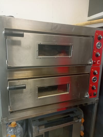 A commercial, two-deck pizza oven is centrally located in an indoor setting. The oven is constructed of stainless steel and features two distinct baking chambers, each equipped with a glass window for monitoring. Both chambers have black, horizontal handles on the left side. The right side of the oven has a red control panel with two knobs, two gauges, and a set of buttons and switches. The oven appears to be in operational condition, though some minor smudges and dust are visible on its surfaces. Beneath the oven, a metal shelving unit is present, holding a large plastic bottle with an orange cap and a black garbage bag. Further down, another appliance, possibly a smaller oven or warming unit, is partially visible. The background consists of a plain, off-white wall. This scene is consistent with a commercial kitchen or food preparation area in a business establishment, such as a pizzeria or restaurant.