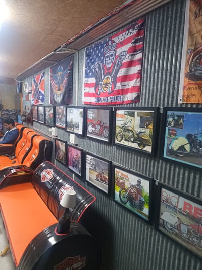 This is an interior shot, likely a themed room or garage, decorated with Harley Davidson and motorcycle-related items. The walls are covered with corrugated metal sheets, on which are hung a Confederate flag, an American flag with a skull design, a map, and various framed motorcycle pictures and advertisements.
In the foreground, there's a custom-made sofa constructed from repurposed black barrels with the "Harley Davidson" logo prominently displayed. The sofa has bright orange cushions. A small lamp is placed on top of one of the barrels, serving as a side table.
In the background, a man is sitting on a similar orange sofa. The room appears to be dimly lit.