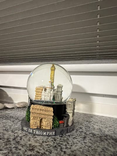 A snow globe with miniature Parisian landmarks sits on a speckled gray countertop. The snow globe features a clear glass dome containing several white and gold buildings, including a tall, slender tower. The base of the snow globe is dark gray and decorated with miniature models of the Arc de Triomphe, a tall ornate building, and green trees. The words "ARC DE TRIOMPHE" are written in white capital letters around the base. In the background, a set of closed gray horizontal blinds covers a window. To the left of the snow globe, a few smooth, light-colored stones are visible on the countertop. The scene is indoors. The image does not provide any information about the city of Sankt Augustin, Germany.