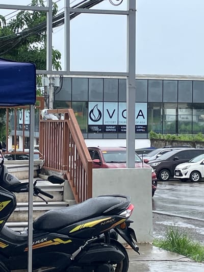The image captures a frontal view of a modern building with large glass windows, displaying the signage "VCA VICTORY CHURCH OF ASIA". Below the church name, service times are listed: "SUNDAY SERVICES 8:30AM 10:30AM 1:30PM 4:30PM". The building is situated in a cityscape, with a street and parked vehicles visible. Among the vehicles are a dark brown SUV, a white sedan, and a red car. In the foreground, a black motorcycle with yellow accents is parked. To the left, a blue awning is partially visible, and a set of outdoor stairs with a brown railing leads up to the building. A few pigeons are perched on the railing of the stairs. The ground appears wet, suggesting recent rain. The overall environment suggests an urban setting in San Jose del Monte, Philippines, with a focus on the church and its immediate surroundings.