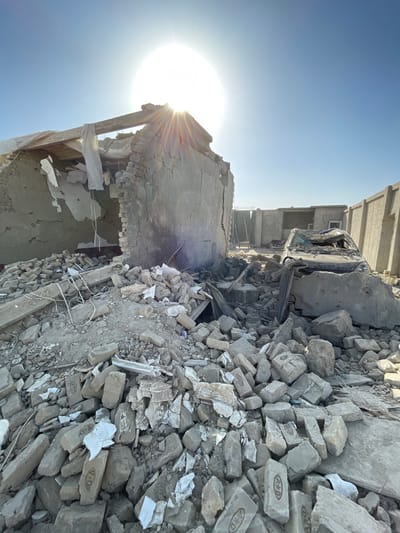 The image displays an extensively damaged urban landscape in Spin Boldak, Afghanistan, under a clear, bright blue sky with a prominent sunburst effect from the high sun. The foreground is filled with a dense accumulation of grey and tan rubble, consisting primarily of fragmented bricks (some marked 'A*D'), broken concrete, and other structural debris, interspersed with pieces of white shredded material. To the left, a partially collapsed masonry building stands, its remaining walls exhibiting cracks, with a portion of its roof missing, revealing exposed wooden beams and draped white sheeting. To the right of this structure, a severely damaged, dark-colored vehicle, resembling a sedan, is observed amidst the rubble; its roof is either caved in or completely absent, and its interior appears compromised and filled with debris. In the background, additional damaged or incomplete structures are visible, along with a tall concrete boundary wall on the far right and a metal staircase ascending in the mid-right portion of the frame. The overall scene appears arid and dusty. No individuals are visible.