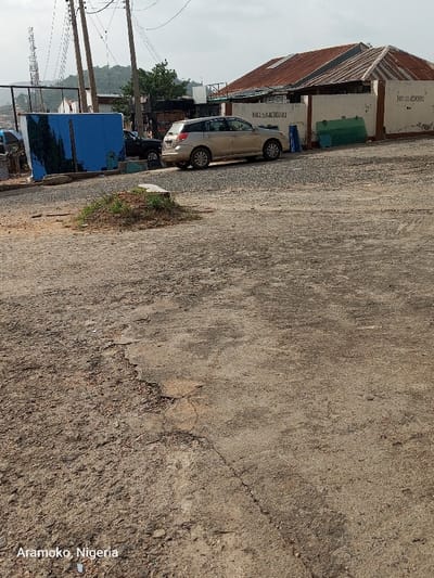 The image shows a cracked, gray asphalt road in the city of Ado Ekiti, Nigeria. It is taken at a slight downward angle. A silver SUV is parked near the edge of the road and is facing the opposite direction of the camera. The vehicle is parked near a building with a red roof. The building appears to be made of concrete with a brown painted wall. A sign on the wall says "No Smoking". There is a large blue structure behind the vehicle and there is a green structure to the right of the vehicle. There is a small pile of dirt and grass in the foreground. There are several electrical poles and power lines in the background. The image appears to have been taken from a person standing on the road. The image is slightly blurry.