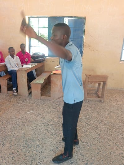 This image depicts a classroom in Mada, Nigeria. A young male with short dark hair, wearing a light blue short-sleeved polo shirt and dark trousers with black slip-on shoes, stands centrally in the frame, facing right and slightly away from the viewer. His arms are raised and blurred, holding an indistinct dark object, suggesting motion. In the background, to the left, two other young males are seated at wooden desk-and-bench units. One wears a white long-sleeved shirt with an emblem and dark trousers, while the other wears a bright pink short-sleeved shirt with an ID badge. A black and red backpack rests on one of the desks. The classroom walls are a light tan color, featuring faint white writing, including partial words like "Light," "Sky," "Stars," and "PALMER." Two windows with blue grates are visible in the background, providing natural light. The floor is covered with mottled grey and light-colored terrazzo tiles. A thin, light-colored stick lies on the floor between the standing individual and the seated students. A small, unadorned wooden table or stool is positioned in the far right background.