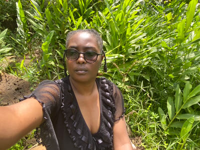 A single individual, an adult female, is depicted in a close-up, selfie-style photograph taken outdoors in Kianyaga, Kenya. She has dark skin and wears dark, round-framed sunglasses. Her hair appears dark and styled in braids or locs. She is dressed in a dark, possibly black, garment with a ruffled V-neckline and sheer, pleated short sleeves. Her right arm is extended, holding the camera. The background is dominated by dense, vibrant green foliage, including broad-leafed plants and various grasses, under bright daylight conditions. A dark, textured object, consistent with rock or bark, is partially visible on the lower left. The specific location within Kianyaga is an outdoor, vegetated area.