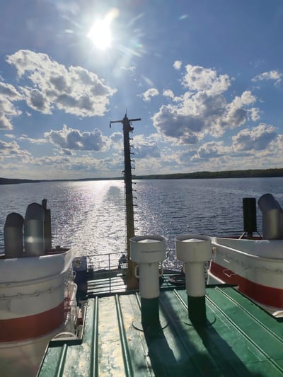 The image depicts a sunny, partly cloudy day on the Volga River in Myshkin, Russia. The view is from the deck of a vessel, looking towards the river. The sun is high in the sky, causing a bright reflection on the water's surface. The river is bordered by tree-lined shores in the distance. The deck of the ship is green and metallic, with various white and red ship components visible, including vents. A tall, metal mast with several attachments stands in the center of the view. The sky is blue with puffy white clouds.