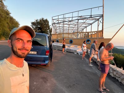 In Svinjišta, Montenegro, a man in a light-colored t-shirt and a baseball cap is taking a selfie in the foreground. Behind him, a blue van is parked, with its driver's side door open. Further back, a group of people are standing on a paved surface near a wooden structure that resembles the hull of a ship. The structure has metal framing and wooden planks, with blue circular decorations. Some of the people are holding drinks or looking at their phones. The environment appears to be outdoors, with trees visible in the background and a clear sky above. The lighting suggests it is late afternoon or early evening, with the sun casting warm light on the scene.