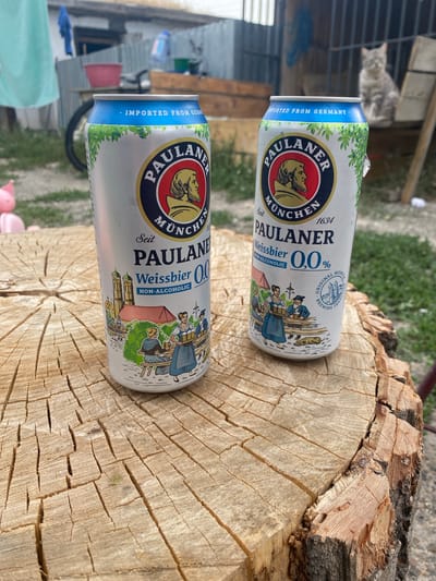 Two cans of Paulaner Weissbier 0.0% are placed on a cross-section of a tree trunk. The can on the left is partially obscured by the can on the right. Both cans are silver with blue and green branding, featuring the Paulaner logo, a depiction of a man's head, and a scene of people in traditional Bavarian attire. The can on the right also shows a circular logo with a depiction of a building and the words "ORIGINAL MUNICH BREWING TRADITION". In the background, there is a blurred scene that includes a fence, some structures, and a cat sitting on a wooden surface. The overall environment appears to be outdoors, possibly in a rural or semi-urban setting.