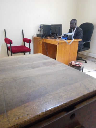 A man in a dark suit and white shirt sits at a wooden desk in an office. He is looking at a computer monitor. Two computer monitors are on the desk, along with a blue and grey bag. To the left of the desk are two red chairs. To the right of the desk is a black office chair. A coiled extension cord with a red reel is on the floor next to the desk. The walls are a pale color. The floor appears to be tiled.
