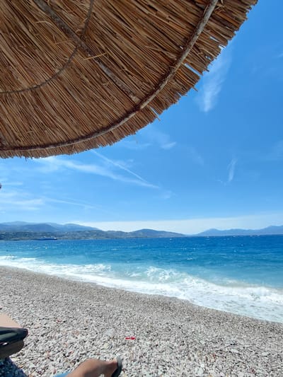 A low-angle, outdoor, full-shot perspective captures a serene beach scene from under a thatched umbrella. The umbrella's woven material dominates the top left of the frame, casting a shadow that emphasizes the bright, clear blue sky above. Wispy white clouds streak across the sky, with one prominent cloud formation in the center. In the distance, a vibrant turquoise sea meets a pebble-strewn beach. Gentle waves crash onto the shore, creating white foam. Beyond the sea, a chain of hazy, green-blue mountains forms the horizon. A large, dark blue and white ferry boat is visible on the water near the mountains. In the foreground, a person's legs and feet are visible, wearing dark sandals, indicating they are relaxing on the beach. The overall impression is one of tranquility and a perfect summer day. The scene is likely the beach of the Municipal Unit of Aegio, Greece, given the characteristic mountainous coastline and clear blue waters of the region.