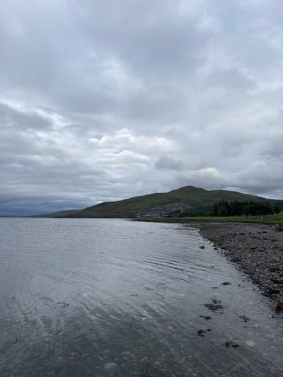 The image captures a vast, serene landscape at Sconser, United Kingdom, characterized by an expansive body of water, a pebbled shore, and a towering mountain under an overcast sky. The foreground features calm water with noticeable ripples, reflecting the muted light of the sky. In the shallower areas near the bank, the water's surface reveals underlying pebbles and darker, irregularly shaped patches, possibly submerged rocks or aquatic growth.

A narrow, dark-toned pebbled and rocky shore separates the water from the land. Beyond this, a large, majestic mountain rises, its slopes densely covered in green vegetation, including grass and clusters of coniferous trees, particularly visible on the lower section. The mountain extends high into the frame, creating a dramatic backdrop.

The sky is heavily overcast with a multitude of grey clouds, varying in density and tone from lighter, diffuse patches to darker, more defined formations. This indicates a cloudy daytime setting with subdued lighting, though no specific time of day like dawn or dusk is suggested. The scene is devoid of human presence or activity, focusing entirely on the natural elements. No text is visible in the image. The overall atmosphere conveys a sense of rugged natural beauty and quiet grandeur characteristic of the Scottish Highlands.